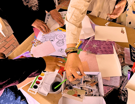 Hands around a craft table with watercolor, collage papers, and art supplies during Vacation Arts