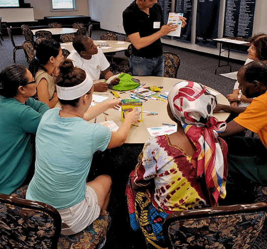BuildaBridge Institute participants around a table making art while a teaching artist holds up a worksheet